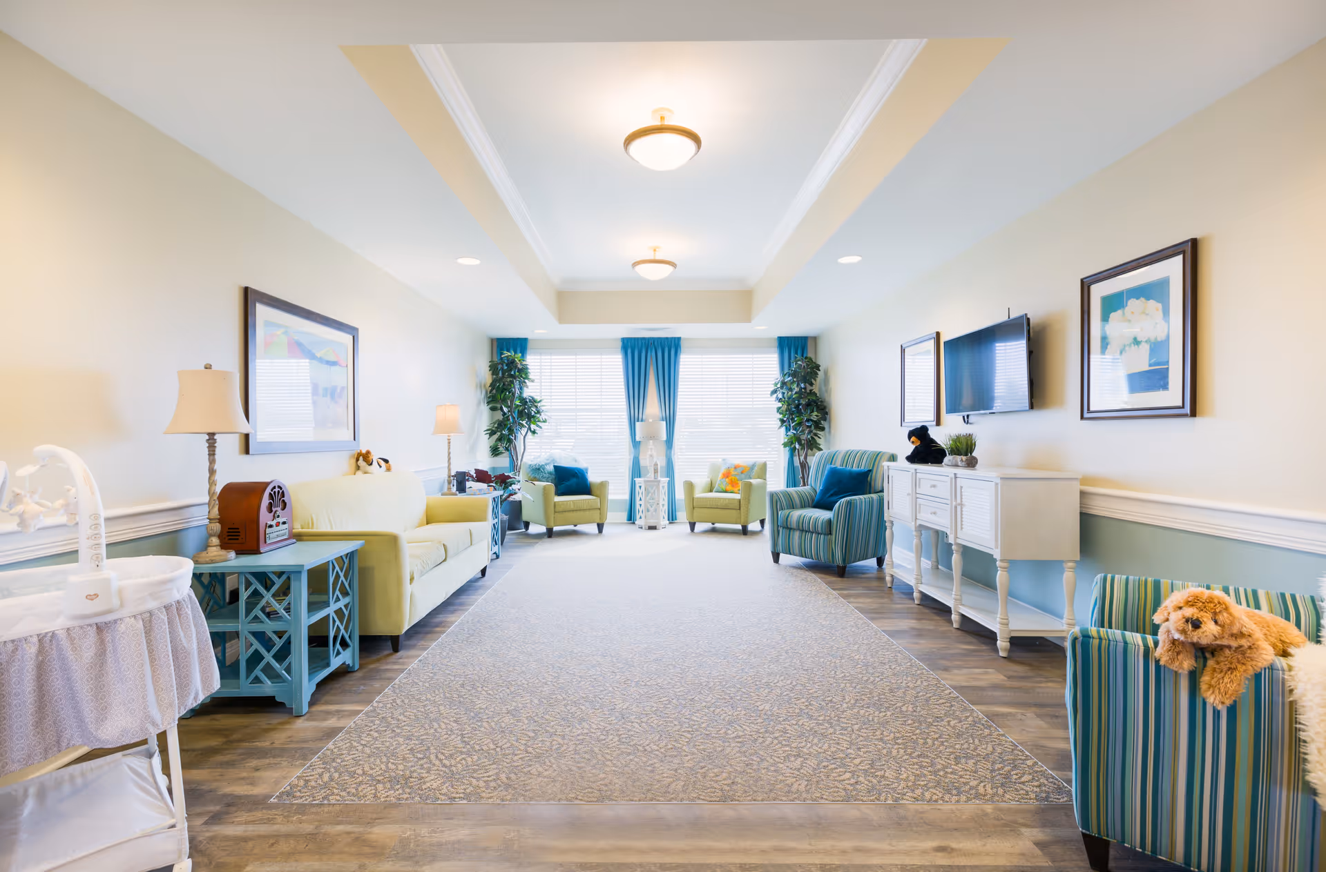 A bright and spacious living room area with a long patterned carpet in the center. The room features a light green sofa, several armchairs in green and blue stripes, side tables with lamps, framed artwork on the walls, a flat-screen TV mounted on the wall, and large windows with blue curtains letting in natural light. There is also a small dog resting on one of the armchairs.