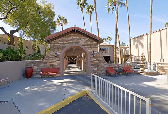 Outdoor courtyard area at Pueblo Norte Senior Living featuring a stone archway with a covered walkway, red cushioned benches on either side, palm trees, and a multi-tiered fountain to the right under a clear blue sky.