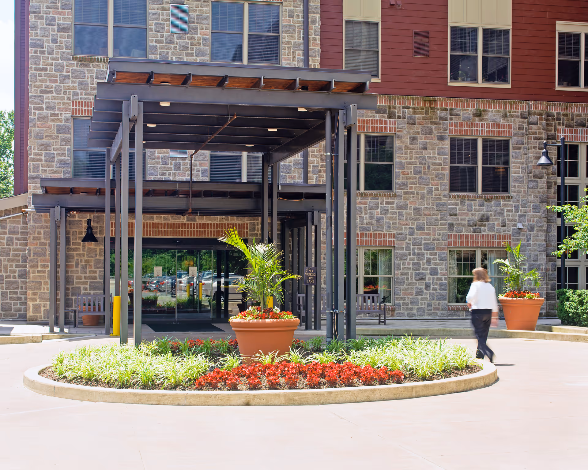 Entrance canopy and circular landscaped driveway with planters in front of a stone-and-brick senior living building, with a person walking by.