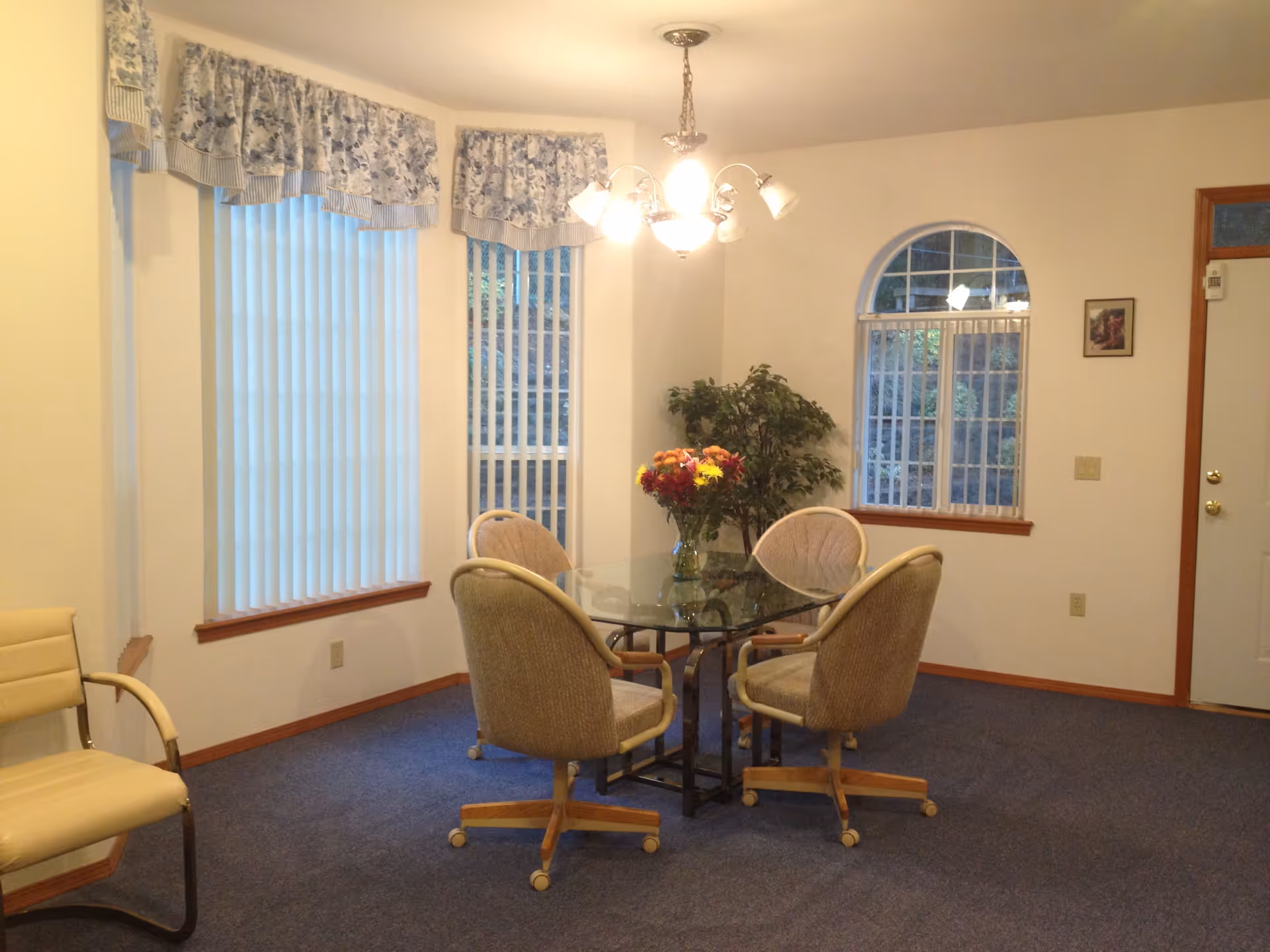 A dining area with a glass-top table surrounded by four beige cushioned chairs on wheels. There is a vase with colorful flowers on the table. The room has blue carpet, white walls, and three windows with vertical blinds and floral valances. A potted plant is in the corner, and a chandelier with multiple lights hangs from the ceiling.