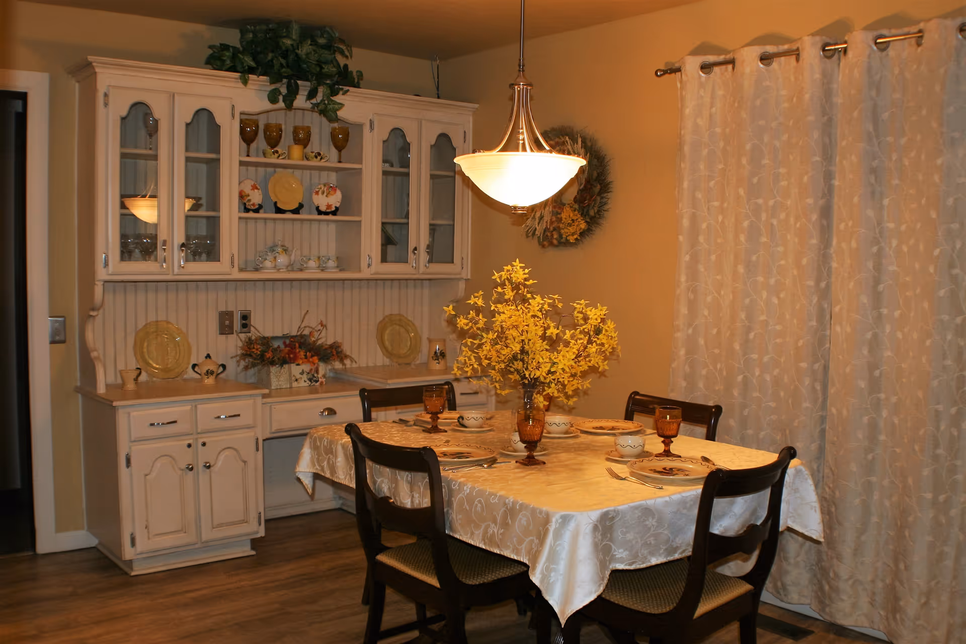 A cozy dining room with a wooden table covered by a cream-colored tablecloth set for four people with plates, cups, and amber-colored glasses. A vase with yellow flowers is in the center of the table. Behind the table is a white wooden hutch displaying decorative plates, cups, and glasses. Beige curtains cover the window, and a hanging light fixture illuminates the room.