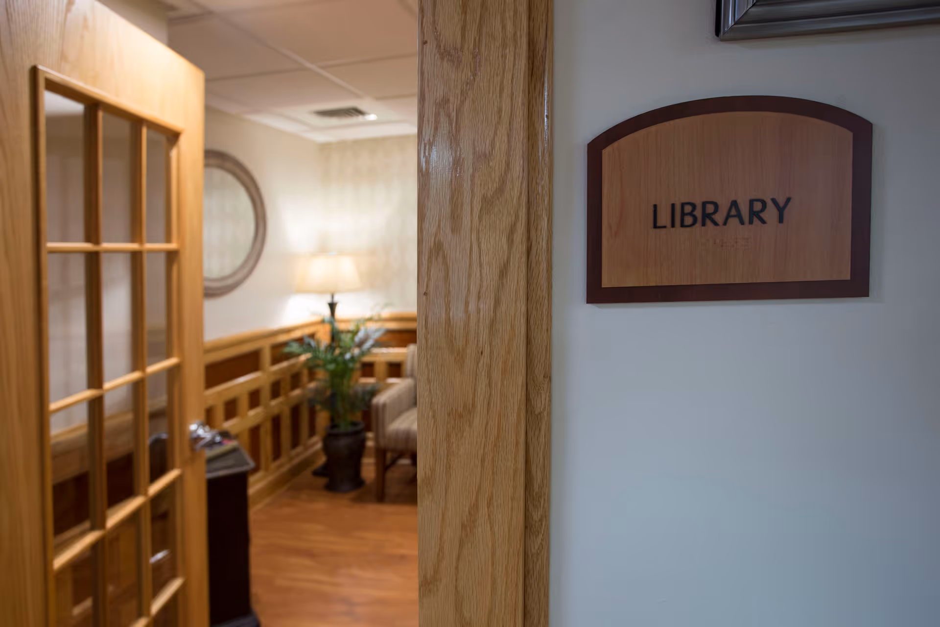 Entrance to a library room in a facility with a wooden door partially open, a wooden sign on the wall reading 'LIBRARY', and a glimpse inside showing a lamp, a round mirror, a plant, and a chair.