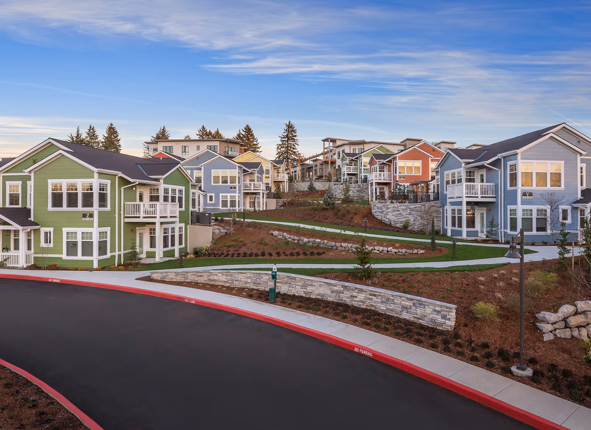 View of a senior living community with multiple two-story houses painted in various colors including green, blue, and orange. The houses are arranged along a curved road with sidewalks and landscaped areas featuring grass, small trees, and stone retaining walls under a partly cloudy sky.