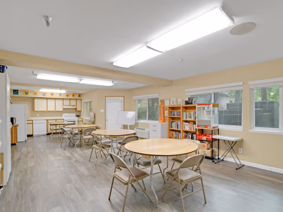 A spacious room with multiple round tables and folding chairs arranged for group seating. The room features a kitchen area with white appliances including a refrigerator, dishwasher, and stove. There are large windows along one wall letting in natural light, a bookshelf filled with books, a popcorn machine, and a whiteboard on a stand near a door. The floor has wood-like vinyl flooring and the walls are painted beige.