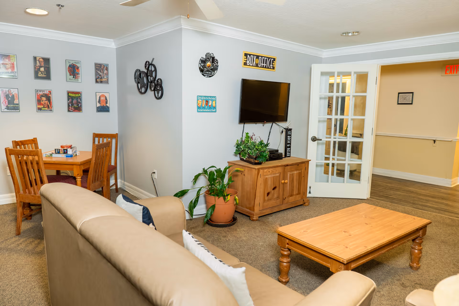 A cozy living room area with a beige couch facing a wooden coffee table and a wooden cabinet with a flat-screen TV mounted above it. The walls are decorated with movie posters and film-themed decor. There is a small wooden table with four chairs in the corner, and a potted plant next to the cabinet. A glass-paneled door leads to a hallway with an exit sign visible.