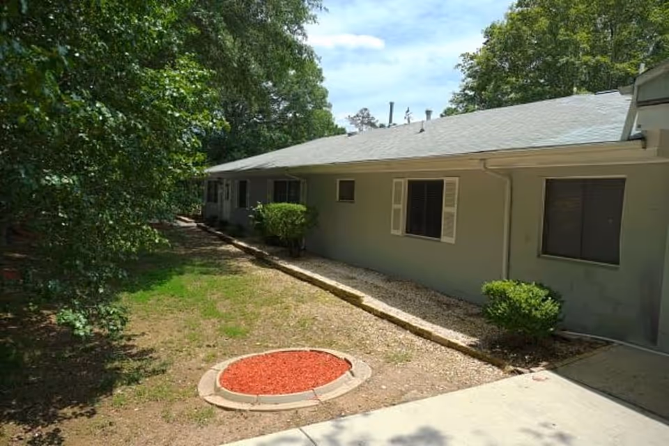 Exterior view of a single-story building with light gray walls and a gray roof, surrounded by trees and bushes. There is a small circular garden bed with red mulch in the foreground and a concrete walkway leading to the building.