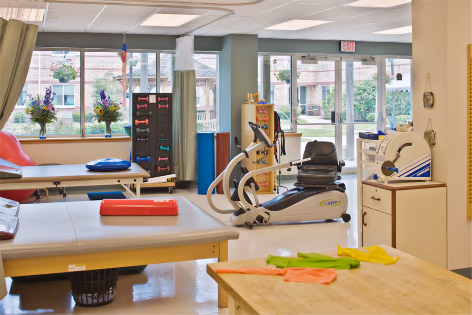 A bright physical therapy or rehabilitation room with exercise equipment including a NuStep recumbent cross trainer, therapy tables, colorful resistance bands on a wooden table, and a rack of small dumbbells. Large windows and glass doors provide a view of an outdoor garden area with hanging flower baskets and a gazebo.