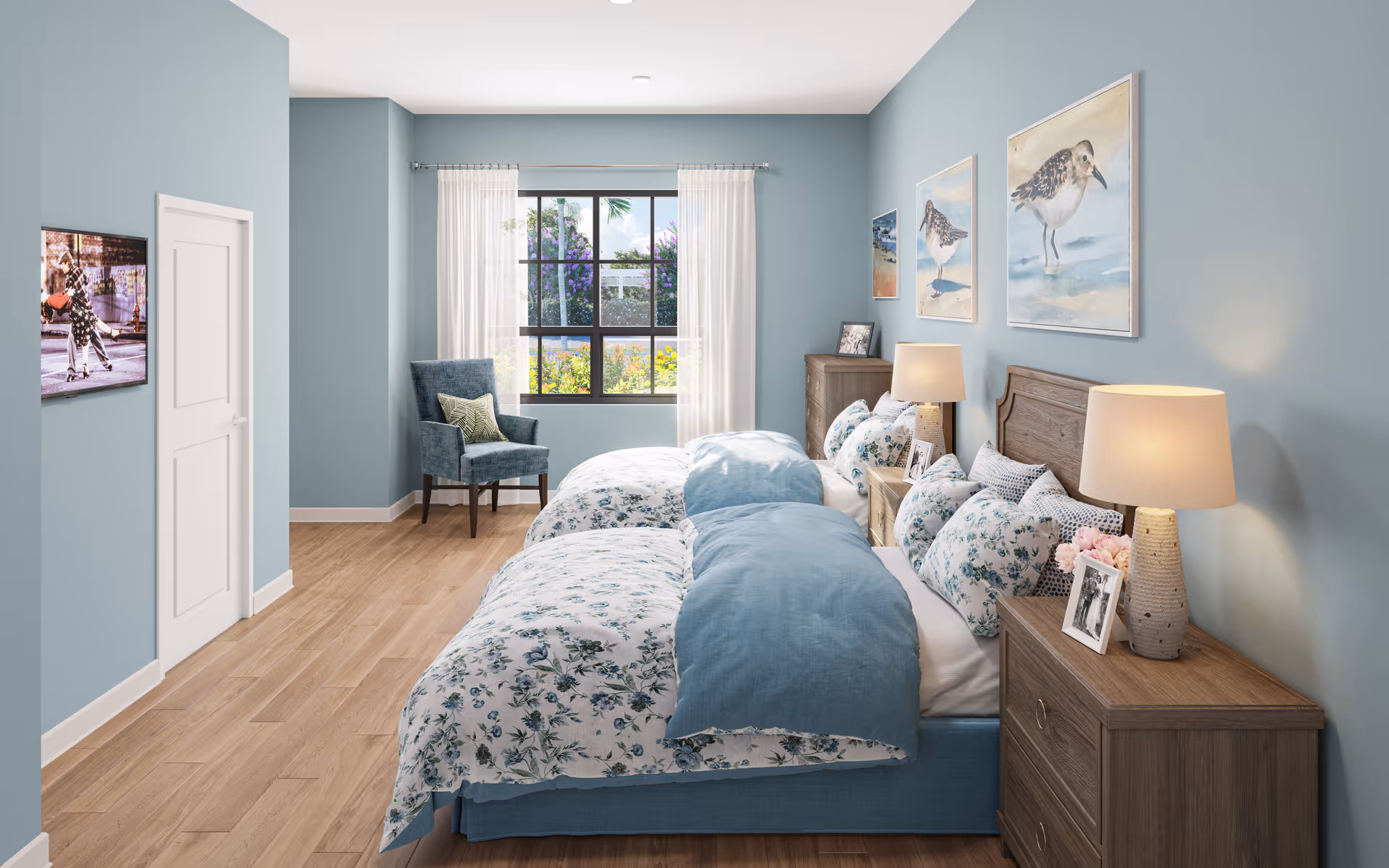 A bright and cozy bedroom with two twin beds featuring floral bedding and blue comforters. The room has light blue walls adorned with bird-themed artwork above the beds. There are two wooden nightstands with lamps and framed photos. A blue upholstered chair with a decorative pillow sits near a window with white curtains, letting in natural light. The floor is wooden, and a closed white door is visible on the left wall.