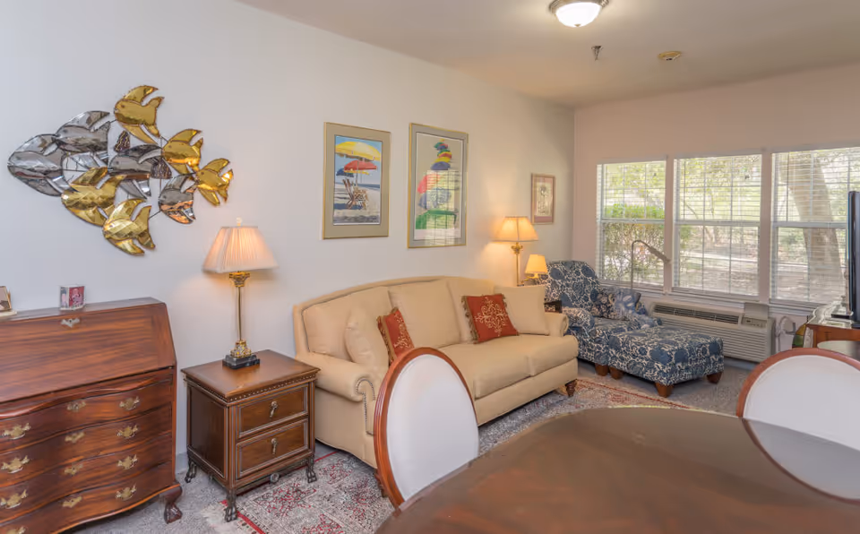 A cozy living room with a beige sofa adorned with red and beige pillows, a blue patterned armchair with matching ottoman, a wooden side table with a lamp, and a wooden chest of drawers. The room has large windows with blinds letting in natural light, framed artwork on the walls, and a decorative metal fish wall art. A round wooden dining table with upholstered chairs is partially visible in the foreground.