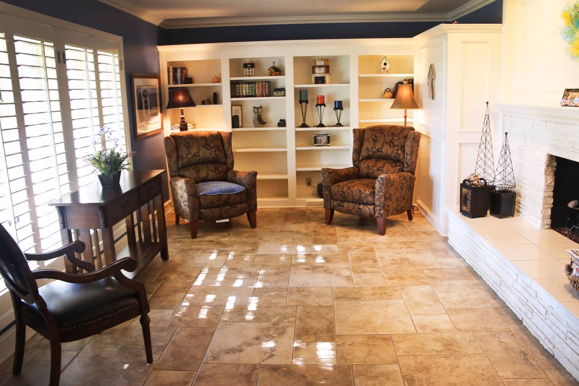 Sunlit sitting area with two upholstered armchairs, built-in white bookshelves, a fireplace, and a small table by shuttered windows.