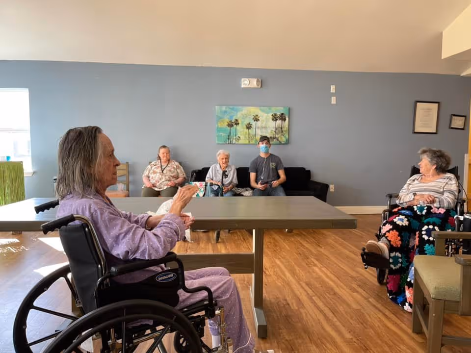 Several elderly residents, including two in wheelchairs, sit around a table in a bright communal living room with blue walls and a palm-tree painting.