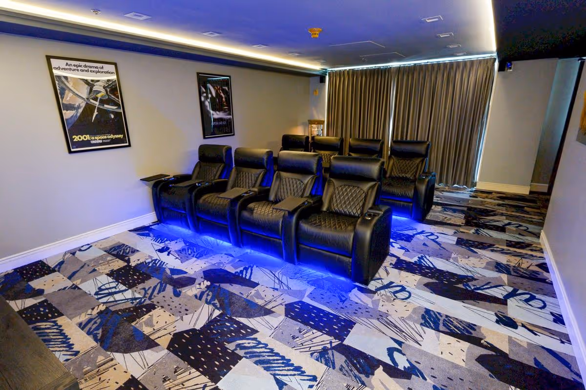 A small home theater room with two rows of black leather recliner chairs illuminated with blue LED lights underneath. The room has patterned carpet flooring and two movie posters on the wall. Heavy curtains cover the window at the back of the room.