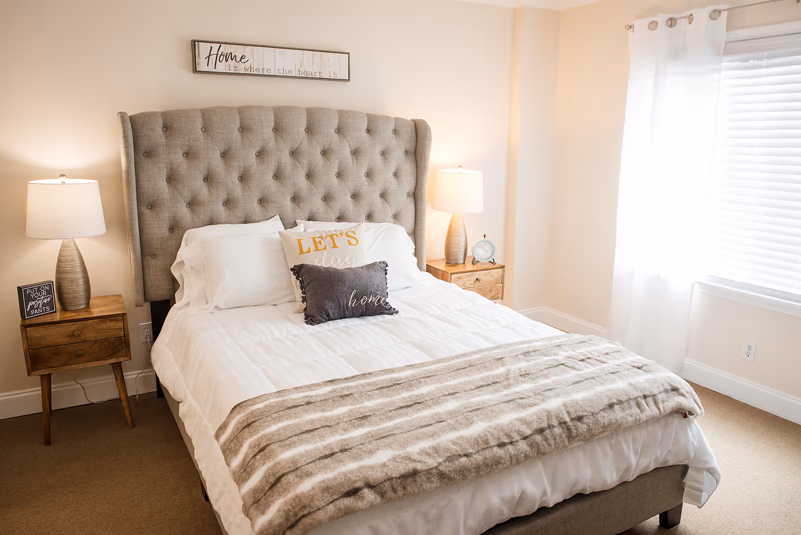 A neatly made bed with a tufted beige headboard in a bright bedroom. The bed has white bedding, two decorative pillows with the words 'LET'S stay' and 'home', and a striped beige throw blanket. On each side of the bed are wooden nightstands with matching lamps. One nightstand has a small sign that reads 'PUT ON YOUR positive PANTS' and the other has a small clock. A window with white curtains and blinds lets in natural light. Above the bed is a wall sign that says 'Home is where the heart is'.