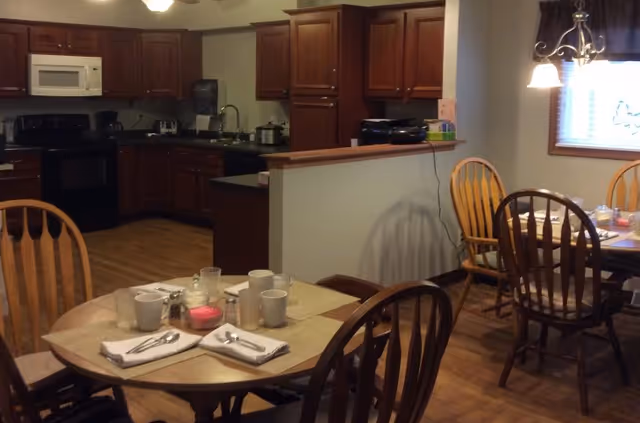 Interior view of a senior living facility dining area and kitchen. The foreground shows a round dining table set with placemats, napkins, cups, and utensils. Wooden chairs surround the table. In the background, there is a kitchen with dark wood cabinets, a microwave, stove, and sink. Another dining table with chairs is visible near a window with blinds and a hanging light fixture.