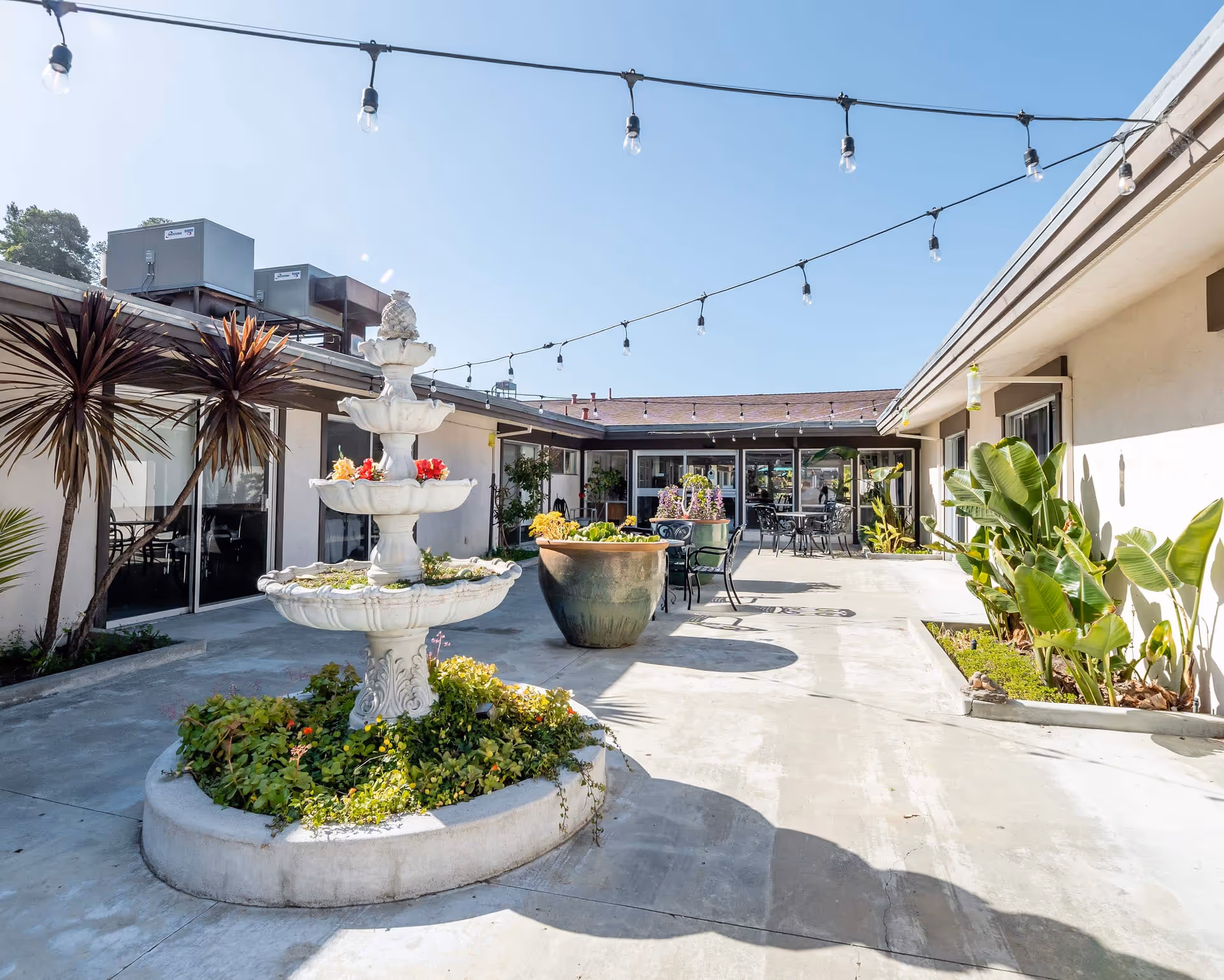 Sunlit courtyard with a tiered fountain, potted plants, string lights, and outdoor seating between single-story buildings.