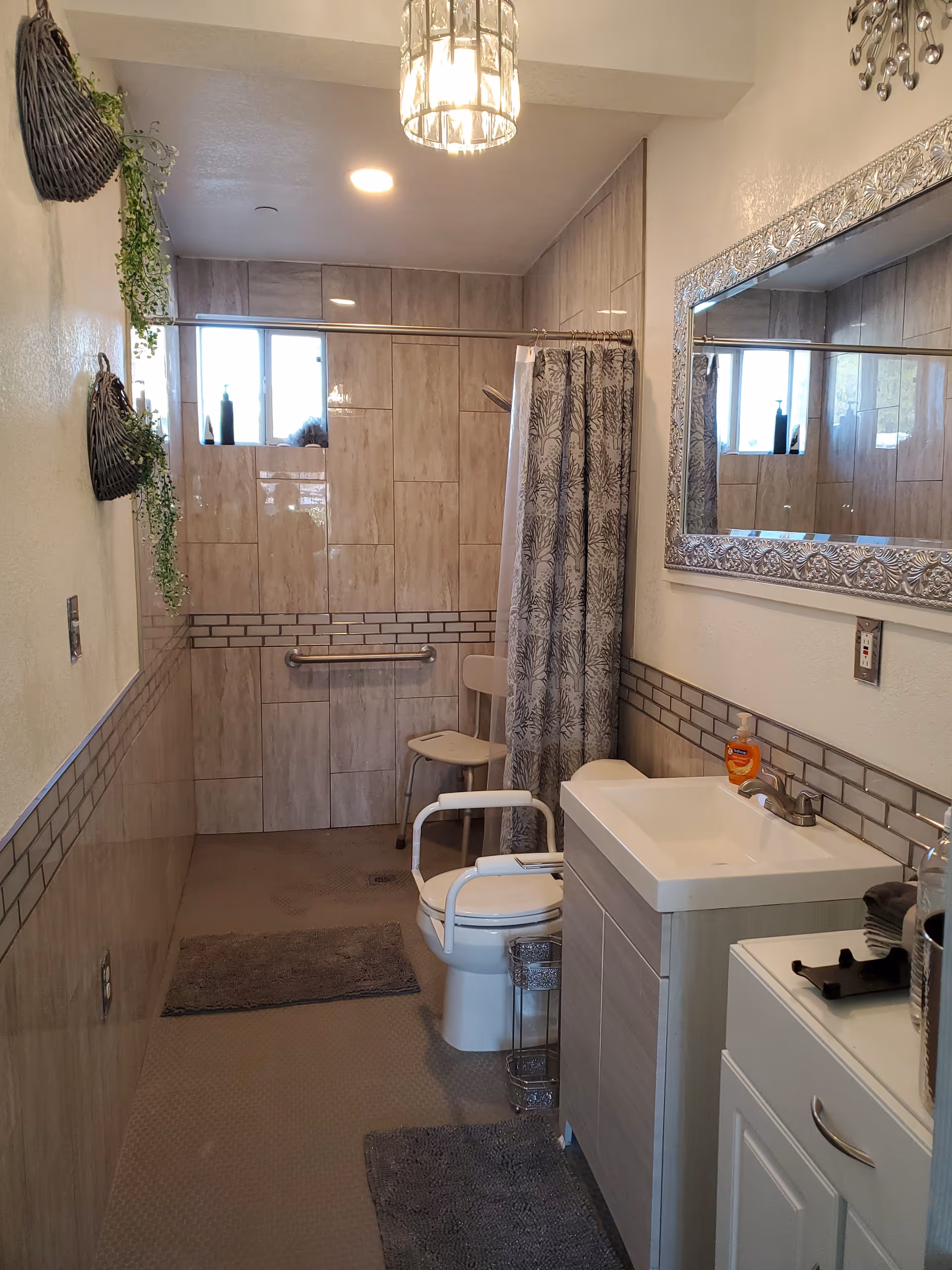 A bathroom with beige tiled walls and floor, featuring a walk-in shower with a curtain, a white toilet with safety rails, a white sink with a cabinet, a large decorative mirror, and hanging plants on the left wall. There are two gray bath mats on the floor and a small chair inside the shower area.