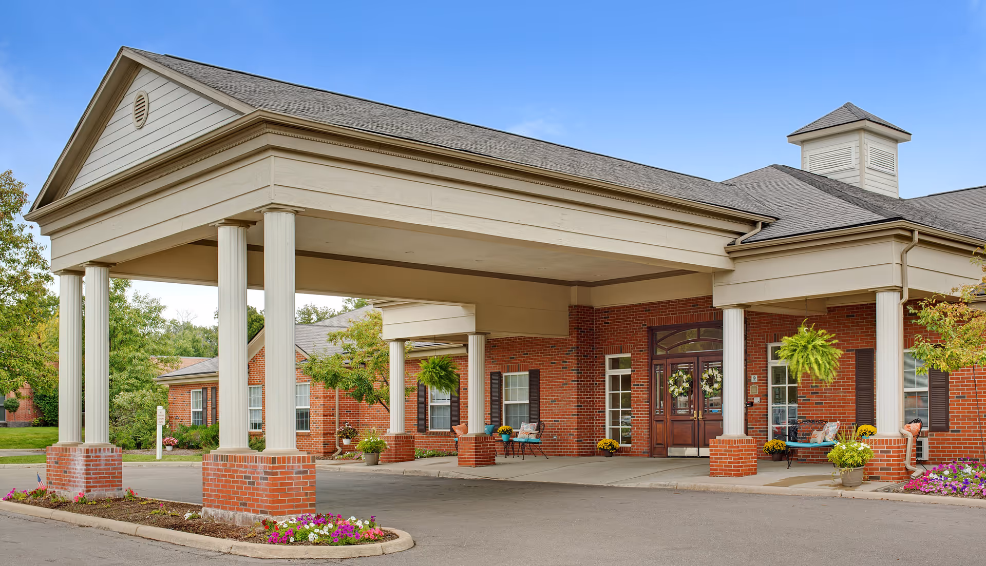 Front exterior view of Downriver Estates Senior Living facility featuring a covered entrance with white columns and brick base, double glass doors, hanging plants, benches, and flower beds under a clear blue sky.