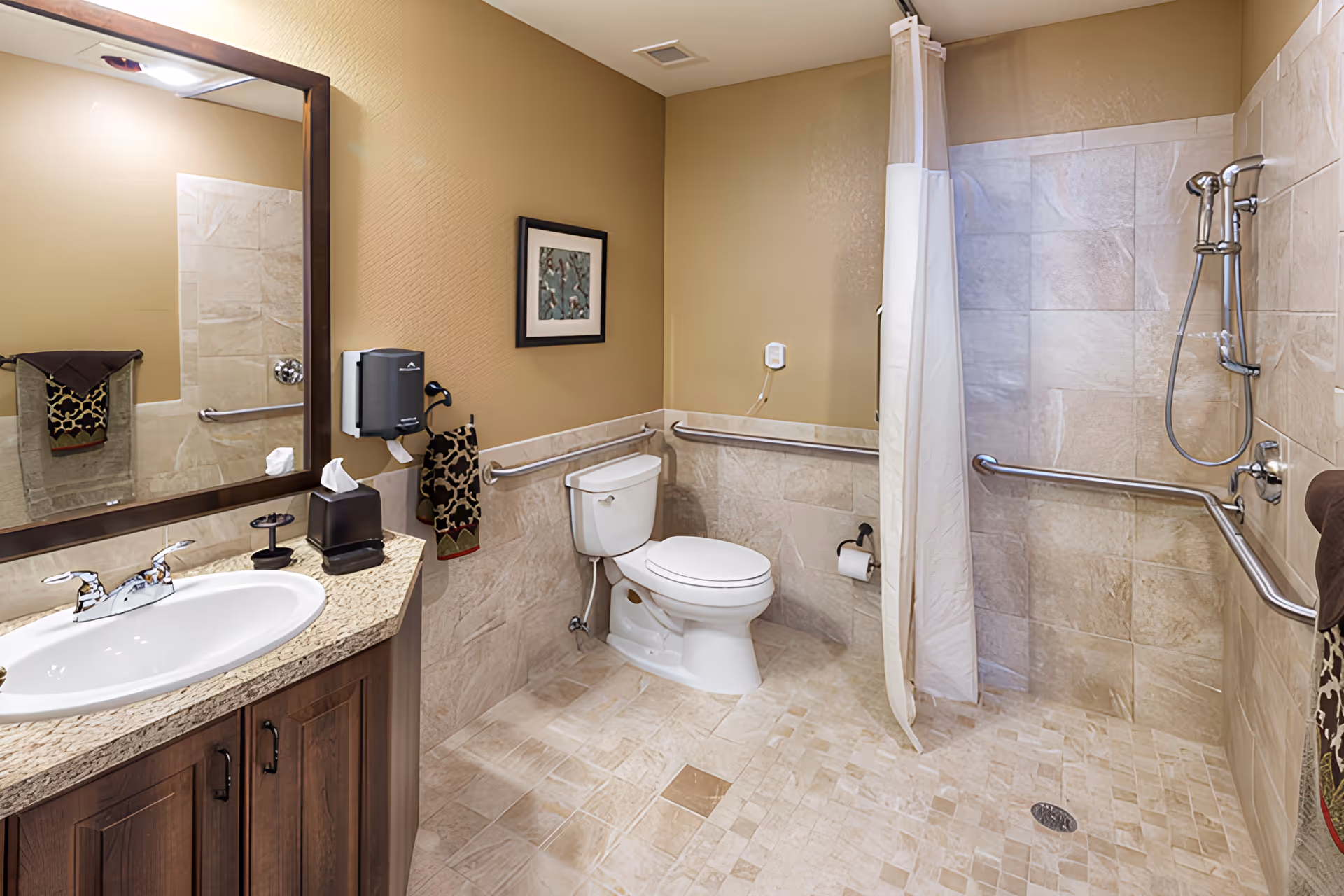 A spacious bathroom with beige tiled floors and walls. It features a white toilet with grab bars on the walls around it, a walk-in shower with a handheld showerhead and grab bars, a white shower curtain, and a wooden vanity with a granite countertop and a sink. A large mirror is mounted above the sink, and there are decorative towels and a framed picture on the wall.