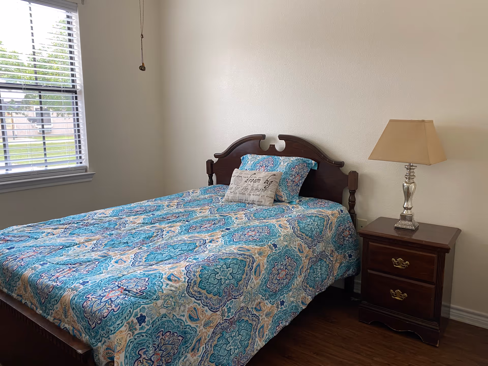 A bedroom with a wooden bed frame and a colorful blue patterned bedspread. There is a pillow with text on the bed and a wooden nightstand with a silver lamp and beige lampshade next to the bed. A window with white blinds is on the left wall, letting in natural light.