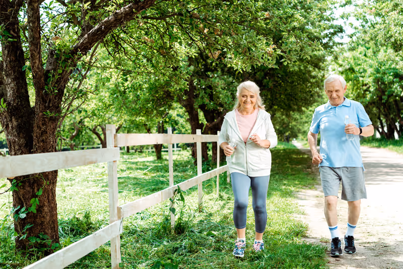An elderly woman and man walking outdoors along a dirt path next to a wooden fence and green trees, both holding water bottles and dressed in casual athletic clothing.
