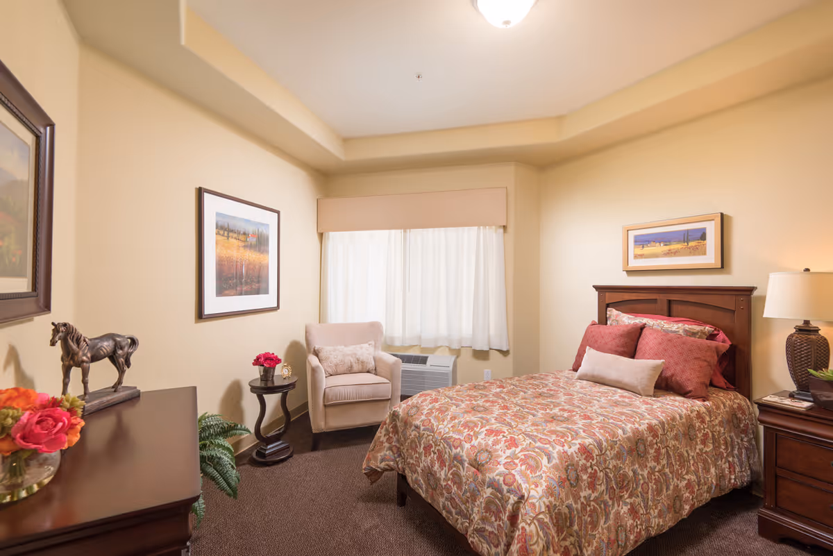 A cozy bedroom with a bed covered in a floral patterned bedspread and multiple pillows. There is a wooden nightstand with a lamp and a small plant, a beige armchair with a decorative pillow, a small round side table with a flower vase, and framed artwork on the walls. The room has beige walls and carpeted floor.
