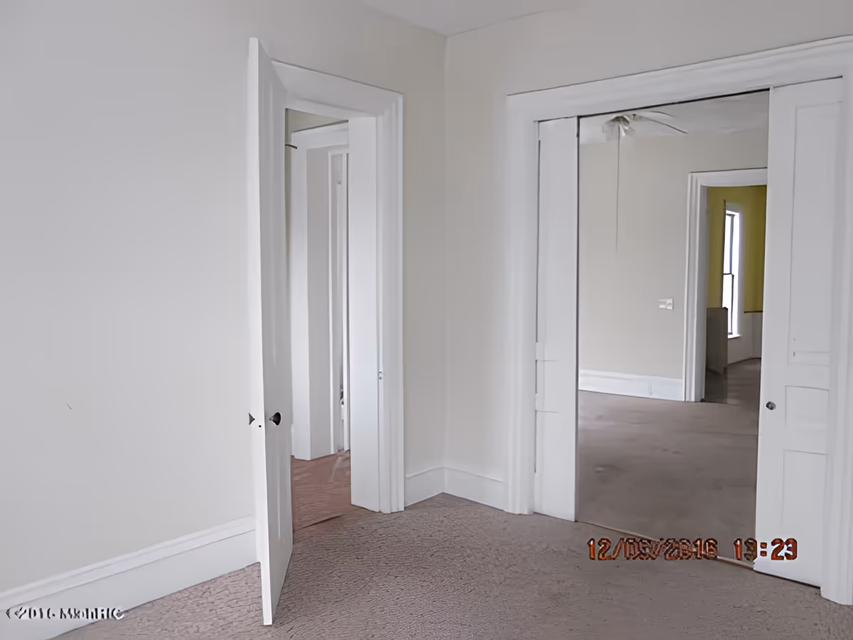 An empty interior room with beige walls and carpeted floor. There are two doorways visible: one with an open door leading to another room, and a second doorway with sliding mirrored closet doors reflecting part of the adjacent room. The adjacent room has a window with yellow curtains.