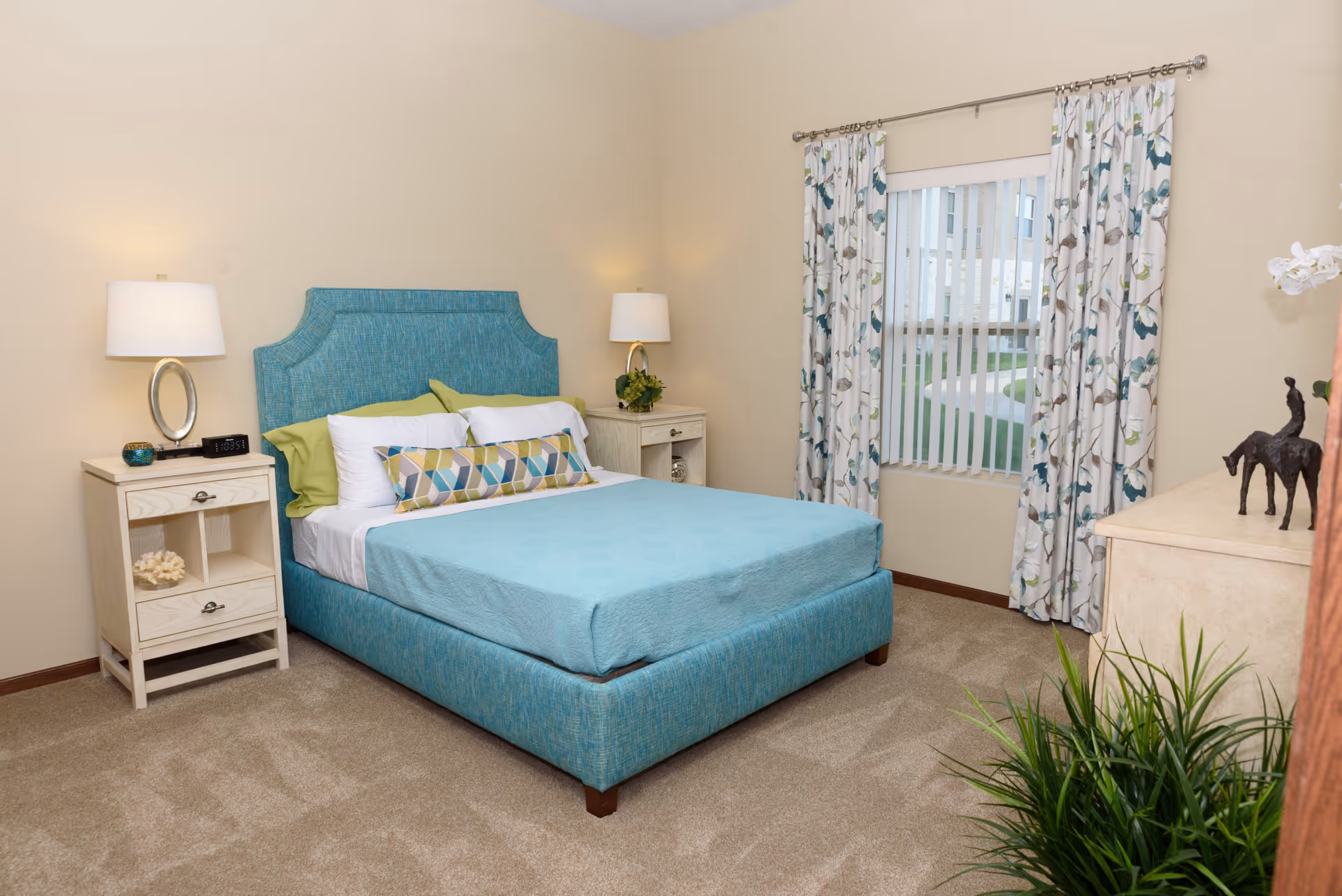 A neatly arranged bedroom featuring a blue upholstered bed with multiple pillows, two white nightstands each with a lamp, a window with floral curtains and vertical blinds, a dresser with decorative items, and a green plant in the corner.