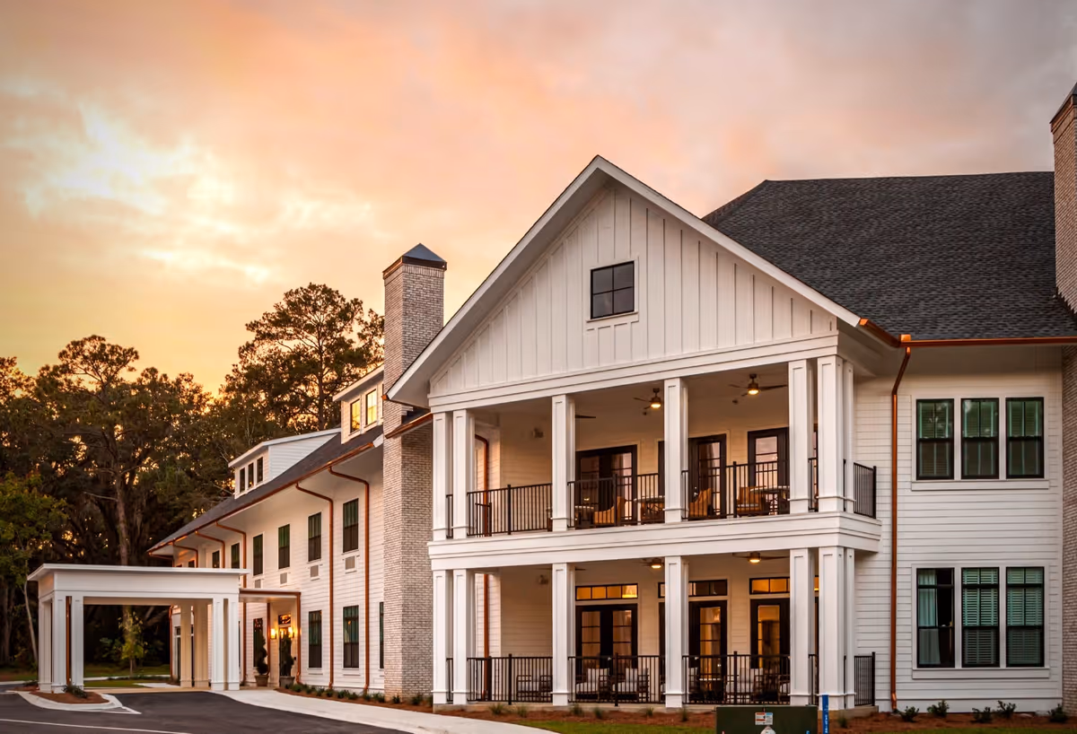 Exterior view of a large, two-story assisted living facility building at sunset. The building features white siding, multiple windows with green shutters, a covered entrance, and a spacious balcony with seating on the second floor. Trees are visible in the background under a partly cloudy sky.