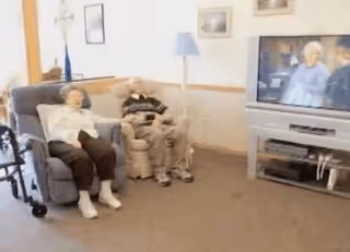 Two people seated in recliners in a common room watching a television.