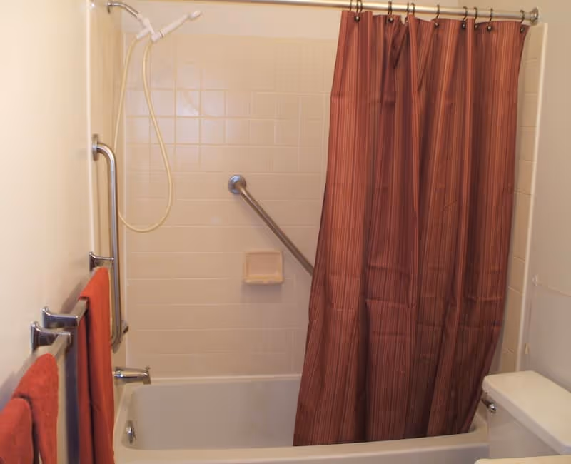 Bathroom with a white bathtub and beige tiled walls. There is a silver grab bar on the wall inside the tub and a red striped shower curtain partially closed. Two red towels hang on a towel rack on the left wall. A white showerhead is mounted above the tub.