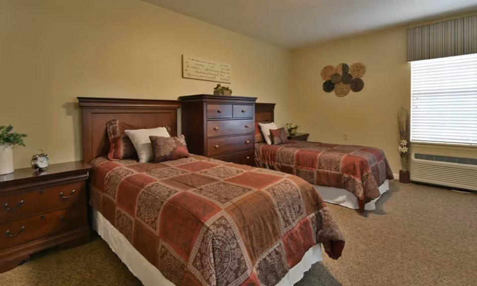 Two twin beds with matching patterned bedspreads, wooden headboards, nightstands and a dresser in a warmly decorated assisted living bedroom with a window.
