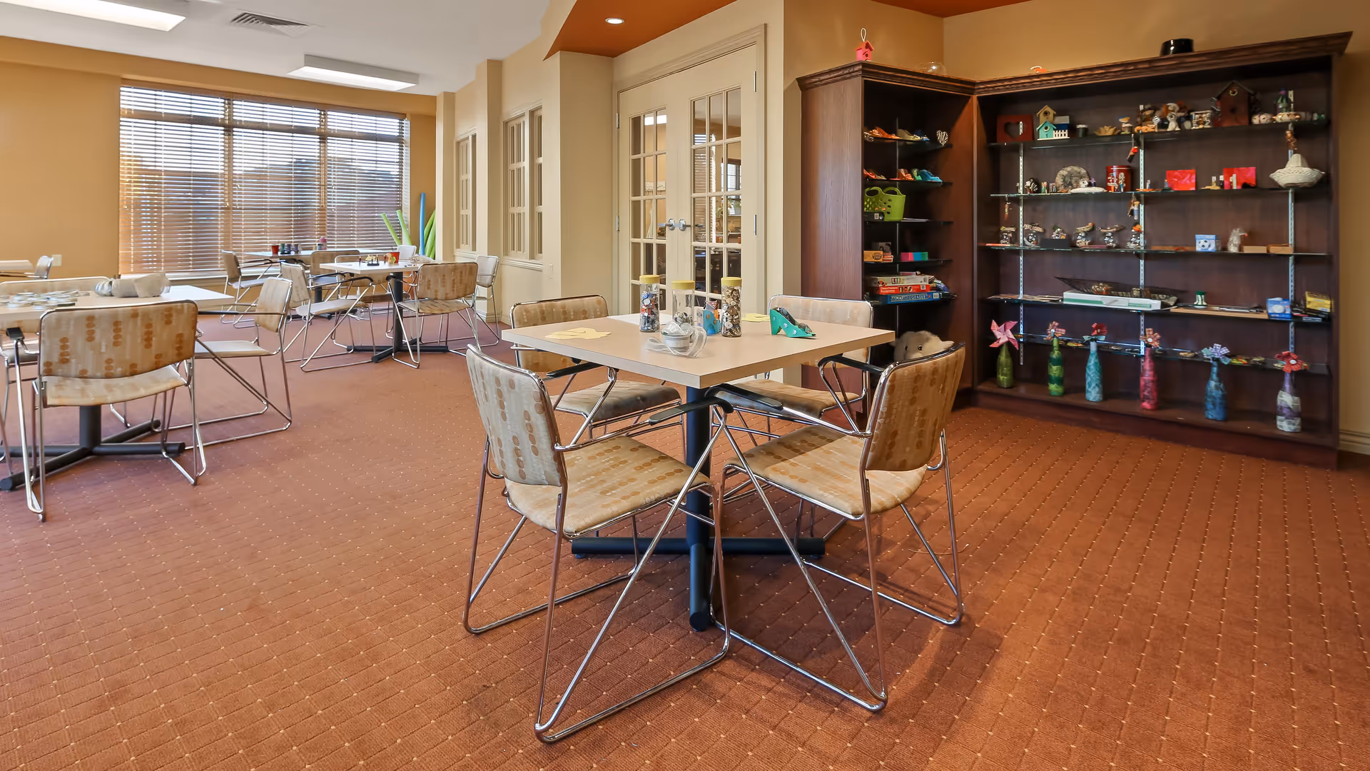 A well-lit room with several square tables and chairs arranged for seating. The room has large windows with blinds allowing natural light to enter. There is a large wooden shelving unit filled with various decorative items and crafts against one wall. The carpet is a warm brown color with a subtle pattern.