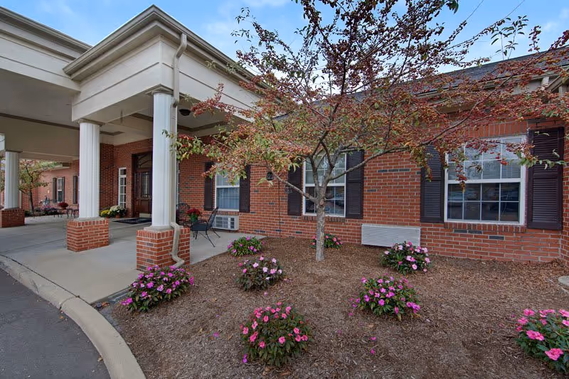 Front entrance of a brick senior living building with a covered portico supported by white columns, windows with shutters, and landscaped beds with pink flowers.