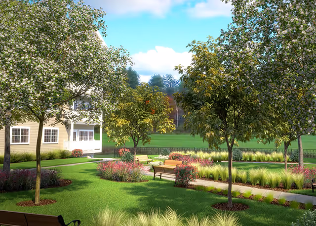 Landscaped courtyard with benches, flowering trees, and a green lawn beside a residential building under a blue sky.