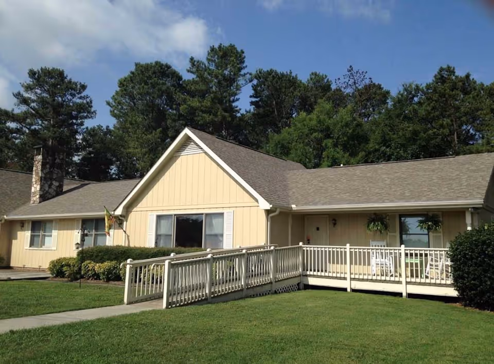 Exterior view of a single-story beige building with a sloped roof, white railings, and a wheelchair accessible ramp leading to the entrance. The building is surrounded by green grass, bushes, and tall trees under a partly cloudy sky.