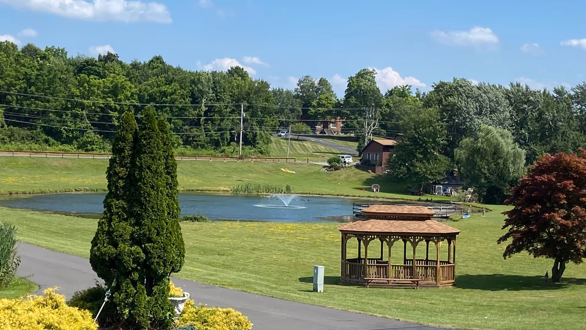 A scenic outdoor area featuring a small pond with a fountain in the center, surrounded by green grass and trees. A wooden gazebo is situated on the lawn near the pond, with a picnic table underneath. The background includes a road, utility poles, and a few buildings partially obscured by trees under a blue sky with scattered clouds.