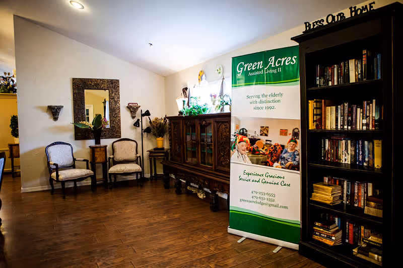Interior of a senior living facility with wooden flooring, two upholstered chairs against a wall with a large decorative mirror above them, a wooden cabinet with glass doors, a tall bookshelf filled with books, and a green and white banner for Green Acres Assisted Living II promoting gracious service and genuine care.