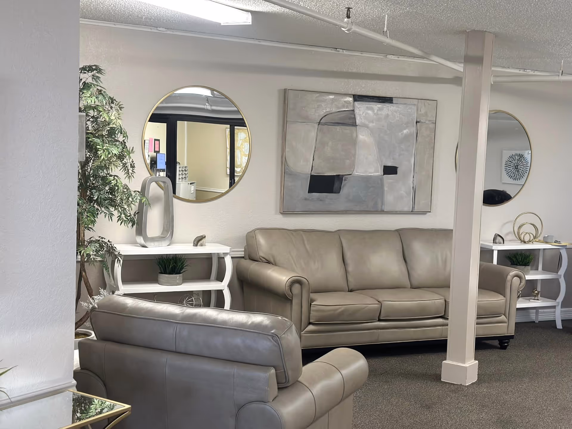 A cozy living room area in a nursing center featuring a beige leather sofa and matching armchair. The walls are decorated with two round mirrors and an abstract painting. There are two white side tables with decorative items and a potted plant in the corner. The room has a neutral color palette and carpeted flooring.