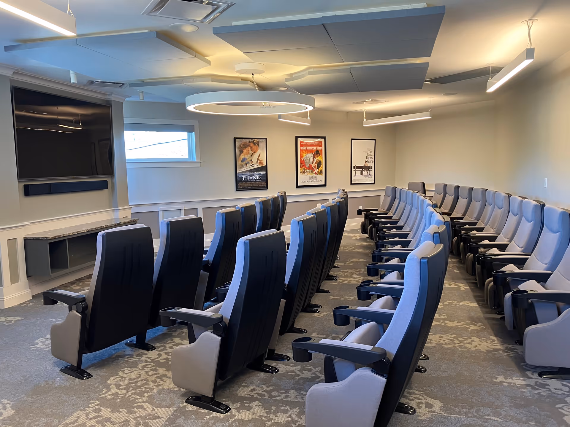 A small theater room with two rows of comfortable, gray and black reclining chairs facing a large flat-screen TV mounted on the wall. The room has a patterned carpet, three framed movie posters on the far wall, and modern ceiling lights.