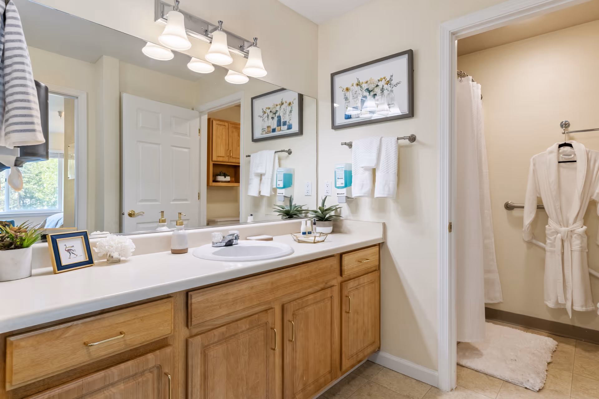 A clean and well-lit bathroom featuring a large mirror above a countertop with a sink. The countertop holds decorative items including a small framed picture, a plant, soap dispensers, and toiletries. Wooden cabinets are below the countertop. On the right, there is a doorway leading to a shower area with a white curtain and a white bathrobe hanging on a hook. Towels are neatly hung on a towel rack beside the mirror.