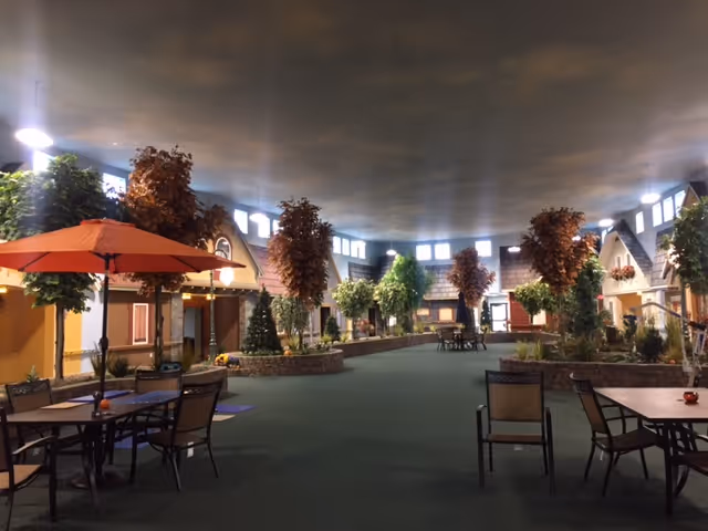 Indoor common area designed to resemble an outdoor village street with artificial trees, small house facades, tables and chairs with umbrellas, and soft lighting from windows near the ceiling.