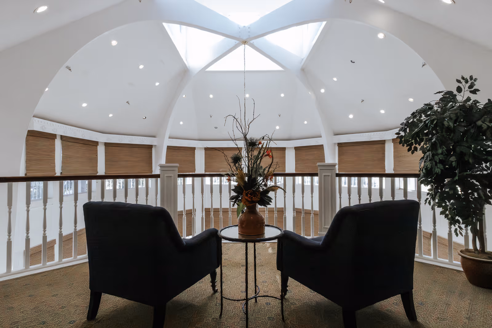 Two dark armchairs facing a small round table with a floral arrangement, situated in a bright, spacious area with a high vaulted ceiling and large windows covered with blinds. A potted plant is placed to the right, and white railings surround the space.