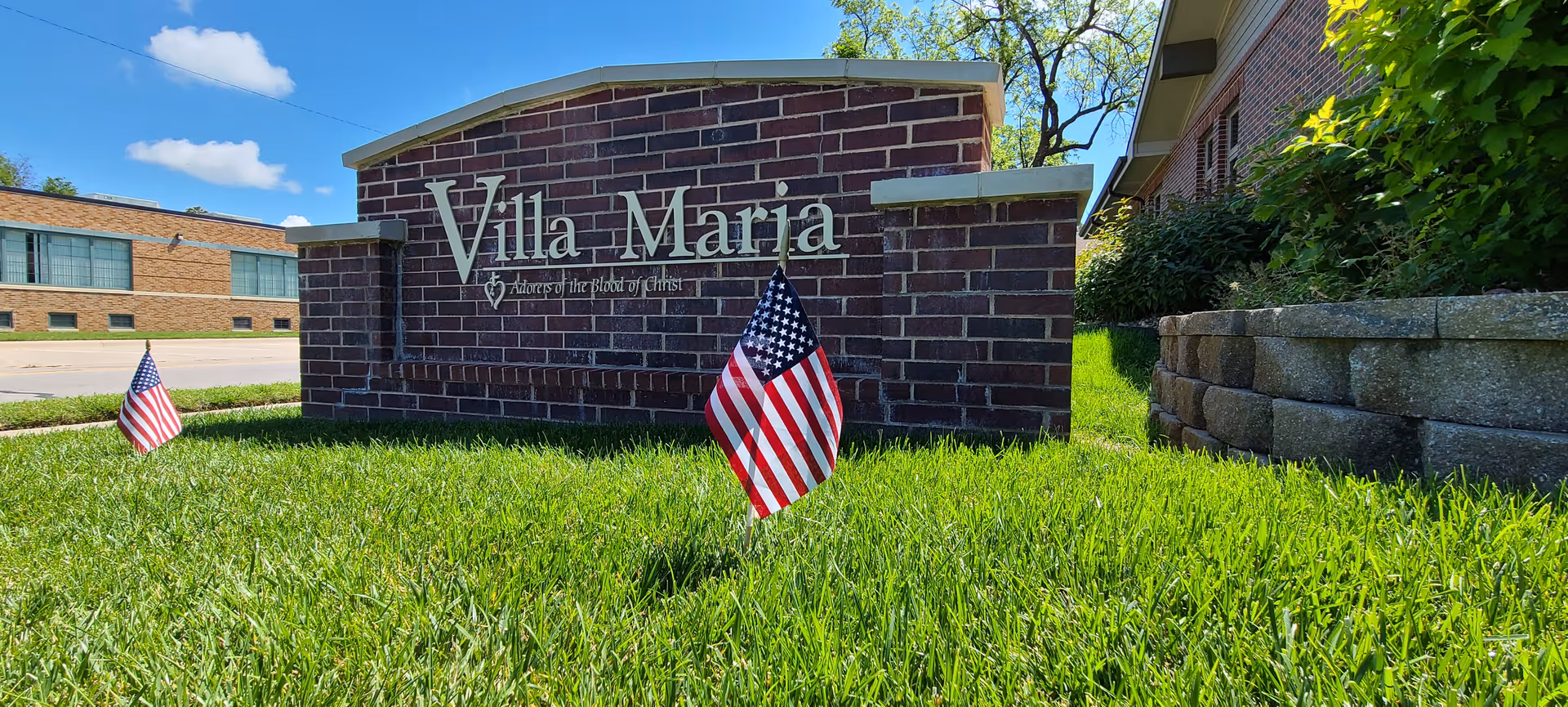 A brick sign for Villa Maria with the phrase 'Advocates of the Blood of Christ' beneath the name. The sign is set in a grassy area with two small American flags planted in the grass. A building and a clear blue sky with some clouds are visible in the background.