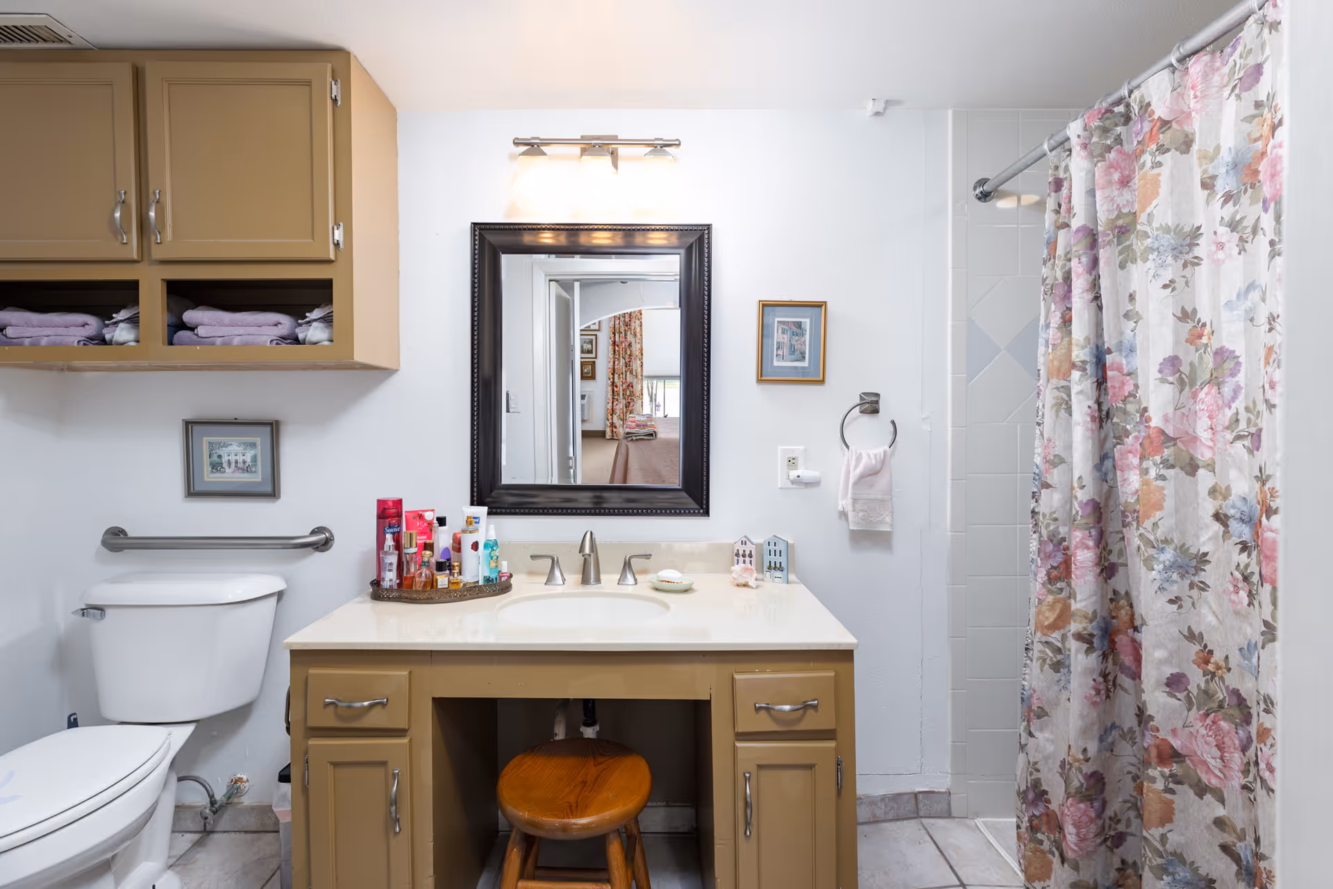 A bathroom with a vanity and mirror, toilet, storage cabinets, and a floral shower curtain.