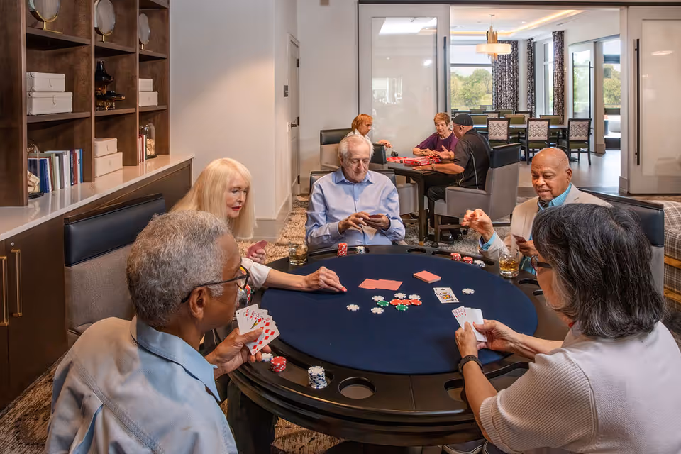 A group of elderly people sitting around a round poker table playing cards and poker chips in a well-lit room with shelves and a large window in the background.
