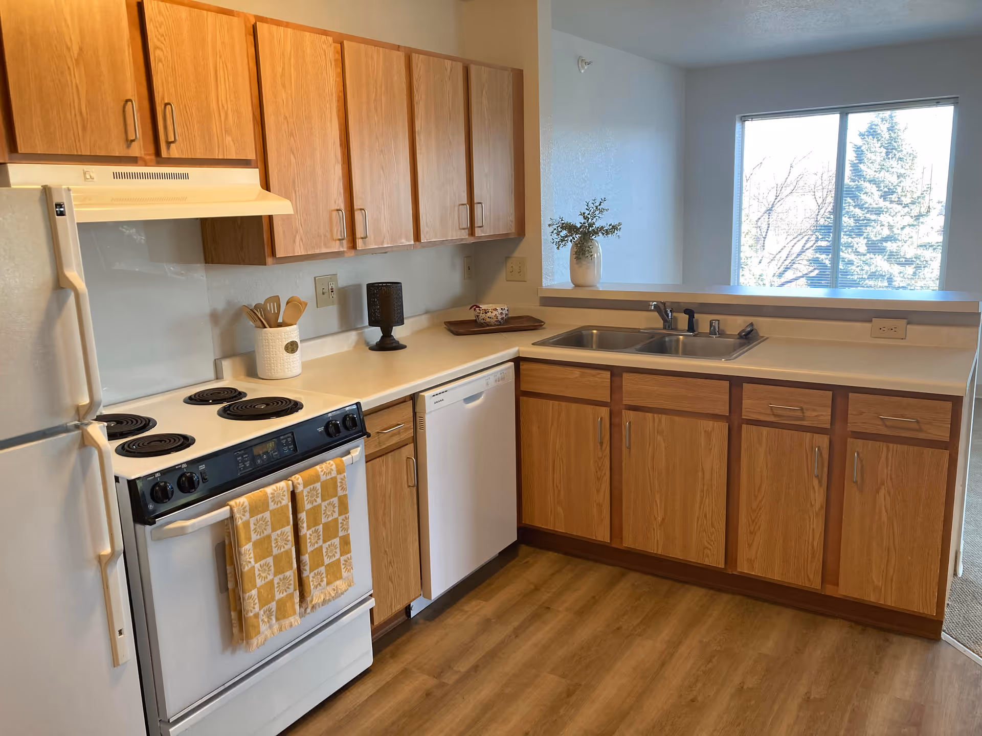 A kitchen with wooden cabinets, a white refrigerator, an electric stove with two yellow patterned towels hanging on the handle, a white dishwasher, a double sink, and a window showing trees outside. The countertop has a utensil holder, a black decorative item, a small bowl on a tray, and a vase with greenery.