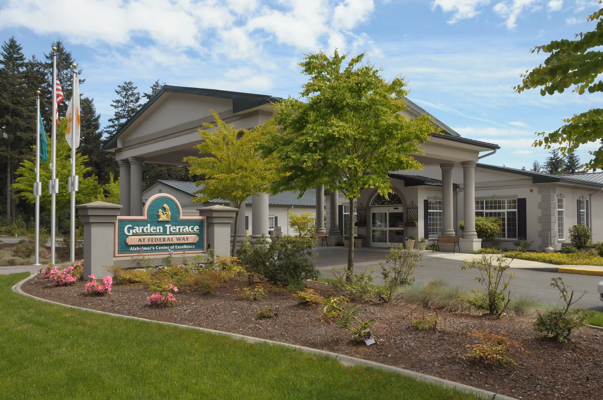 Exterior view of Garden Terrace Healthcare Center of Federal Way showing the entrance with a covered driveway, columns, and surrounding landscaping including trees, bushes, and flowers. There are three flagpoles with flags near the entrance sign.