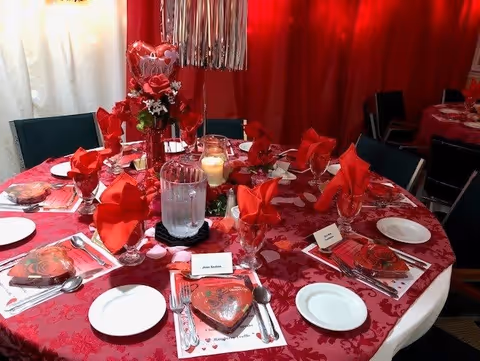 A round dining table set for a special occasion with red and white decorations, including red napkins folded in glasses, a heart-shaped box of chocolates at each place setting, a centerpiece with red roses and a candle, and place cards with names. The background features red curtains.