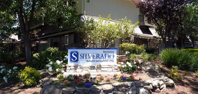 Outdoor view of Silverado Berkeley Memory Care Community sign surrounded by landscaped garden with flowers, bushes, and trees in front of a residential-style building.