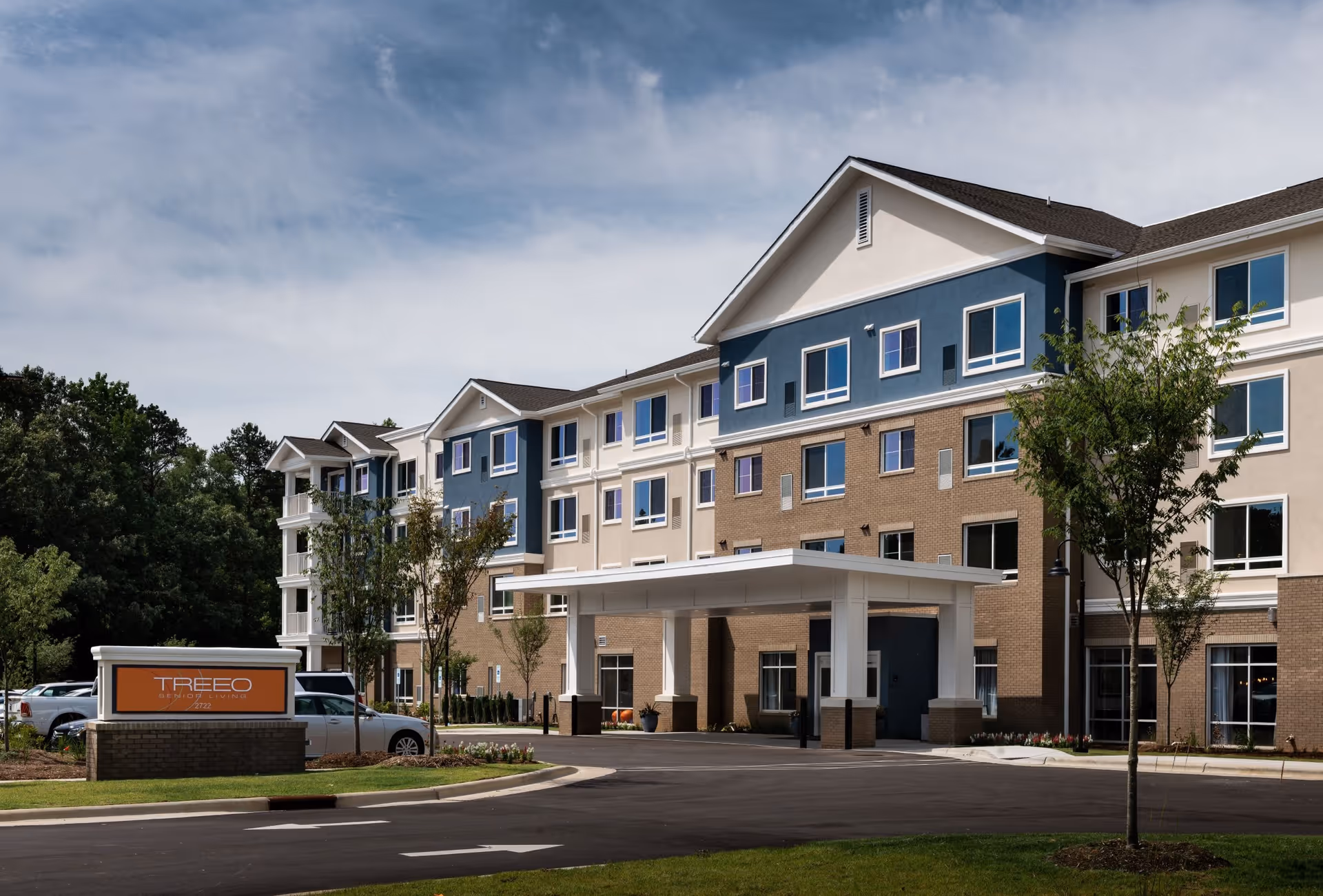 Exterior view of a modern, multi-story senior living facility with a covered entrance, several windows, and a parking area. There are small trees and landscaped areas around the building under a partly cloudy sky.